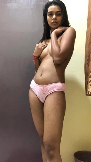 Woman with dark hair in pink underwear posing indoors, hand on chest.