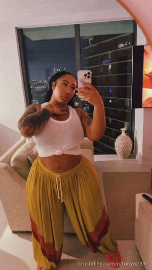 Woman with tattoos in white crop top and yellow pants takes mirror selfie in modern living room.