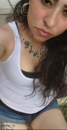 Woman with dark hair and neck tattoo wearing white tank top and denim shorts outdoors.