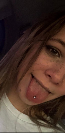 Woman with freckles and tongue piercing, wearing white top, close-up selfie.