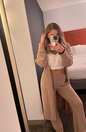 Woman in beige cardigan and white crop top takes mirror selfie in hotel room.