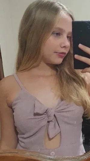 Woman with long blonde hair in a light purple top with bow detail, taking mirror selfie indoors.