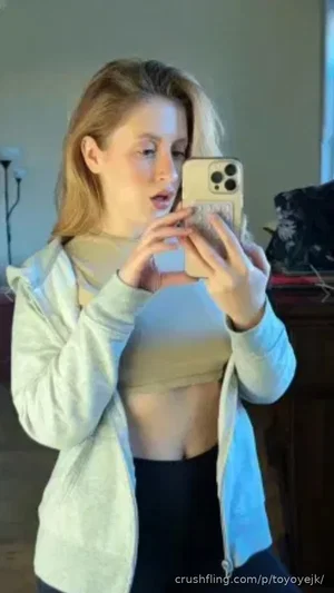 Woman with blonde hair in a light hoodie and crop top, taking mirror selfie indoors.