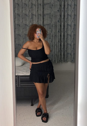 Woman with curly hair taking mirror selfie in bedroom wearing black outfit.