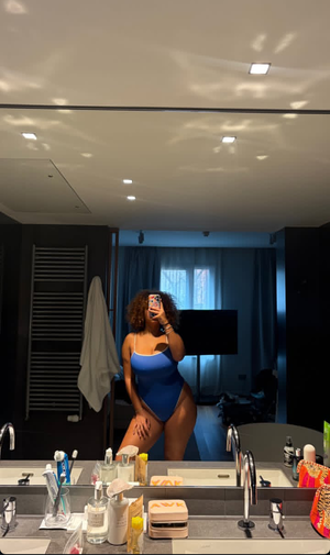 Woman with curly hair in blue swimsuit taking mirror selfie in bathroom.