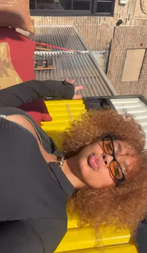 Curly-haired woman with glasses in black top posing outdoors.