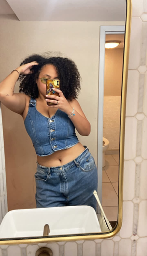 Woman with curly hair taking mirror selfie in bathroom wearing denim outfit.