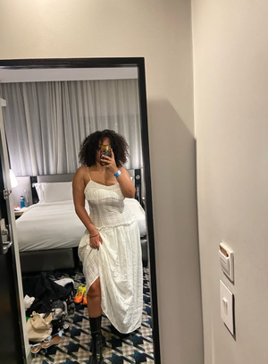 Woman with curly hair in white dress taking mirror selfie in bedroom.