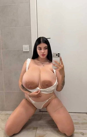 Woman with dark hair kneeling in bathroom, wearing white lingerie, holding phone for mirror selfie.