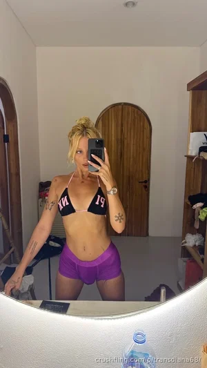 Woman in black and pink bikini top and purple shorts takes mirror selfie in bedroom.