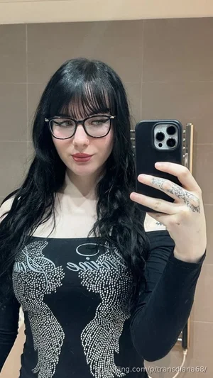 Woman with long black hair and glasses takes mirror selfie in black embellished top.