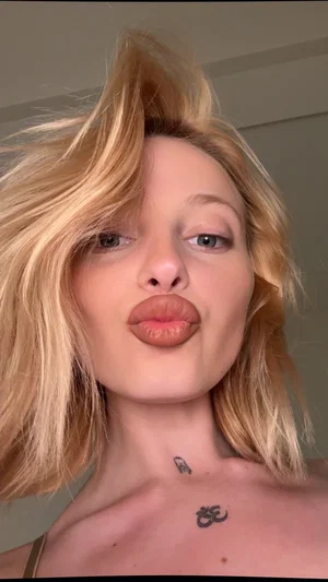 Blonde woman with tattoos on collarbone, topless, making a kiss face indoors.