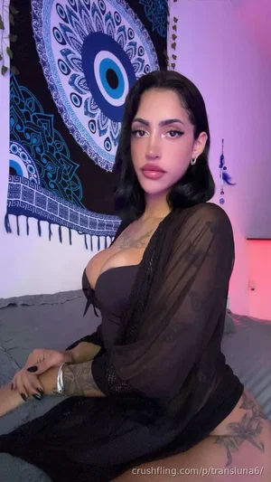 Woman with dark hair and tattoos wears black sheer top, sitting on bed with mandala tapestry behind.