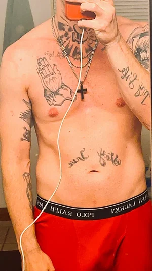 Shirtless man with tattoos, wearing red pants, holding a drink indoors.