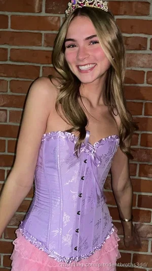 Woman in purple corset and pink skirt with tiara, smiling in front of brick wall.