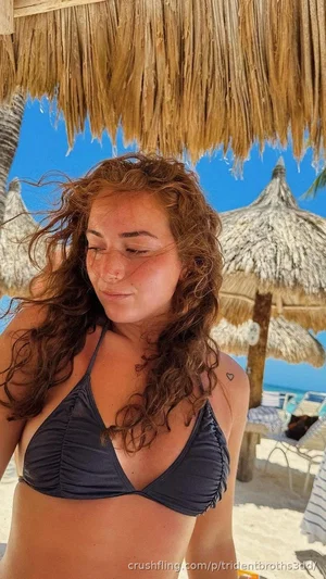 Woman with curly hair in a dark bikini top under a thatched roof at a beach.