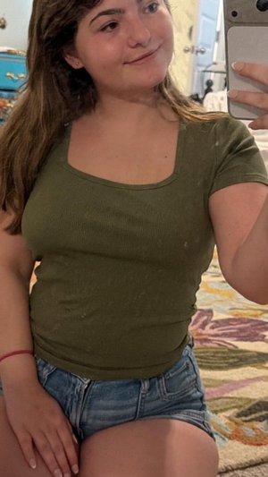 Curvy woman in green top and denim shorts taking selfie indoors.