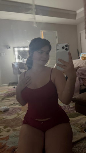 Woman in red bodysuit poses for mirror selfie in bedroom.