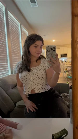 Woman in white patterned top and black pants takes mirror selfie in living room.