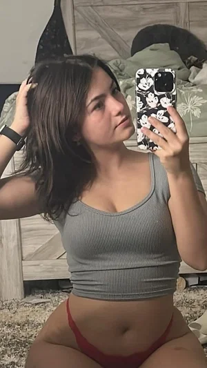 Woman in gray crop top and red underwear posing in bedroom mirror selfie.