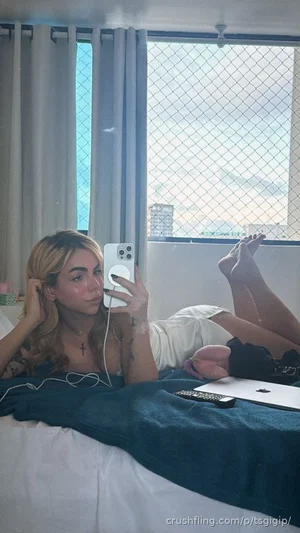 Woman with blonde hair lying on bed, wearing white top, holding phone for mirror selfie.