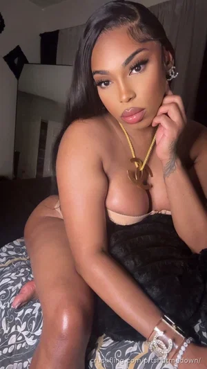 Woman with long dark hair wearing black fur top and beige bra, sitting on patterned bed in bedroom.