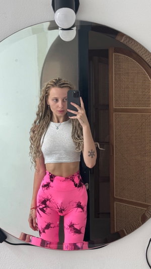 Woman with curly hair in white crop top and pink patterned pants taking mirror selfie indoors.