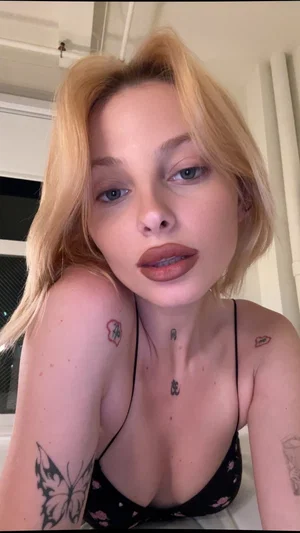 Blonde woman in black floral bra with tattoos on arms and chest, indoors.