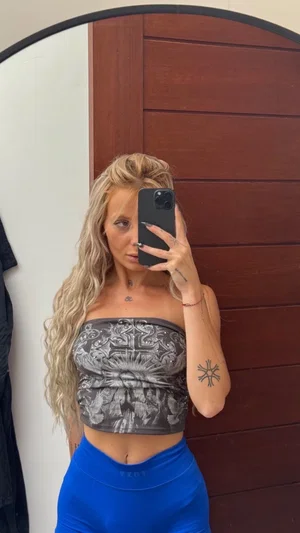 Blonde woman in strapless top and blue leggings takes mirror selfie indoors.