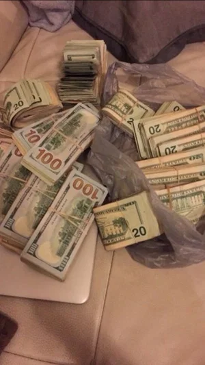 Stacks of US dollar bills on a bed with plastic bags and a laptop.
