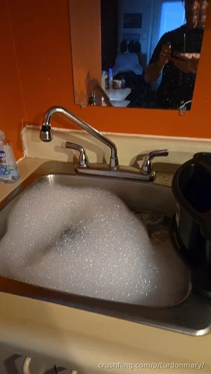 Person in dark shirt taking mirror selfie in bathroom with foamy sink.