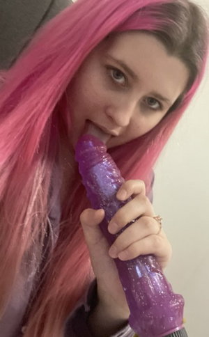 Pink-haired woman with purple dildo in mouth indoors.