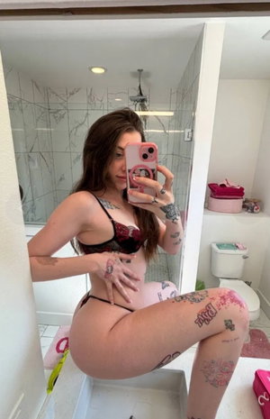 Tattooed woman in lingerie taking mirror selfie in bathroom.