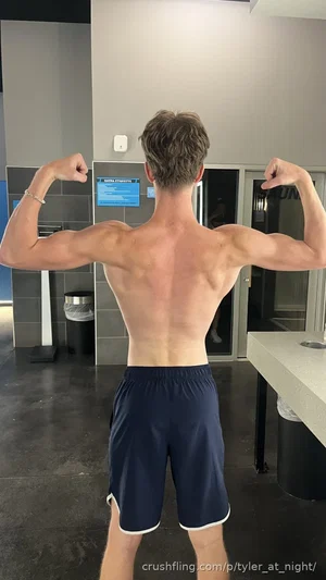 Shirtless man flexing back muscles in gym, wearing navy shorts.
