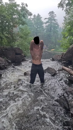 Shirtless man standing in a rocky stream surrounded by trees, wearing dark pants.