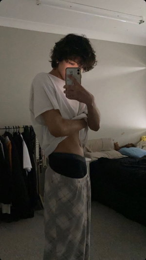 Person with curly hair in white t-shirt and patterned pants, taking mirror selfie in bedroom.