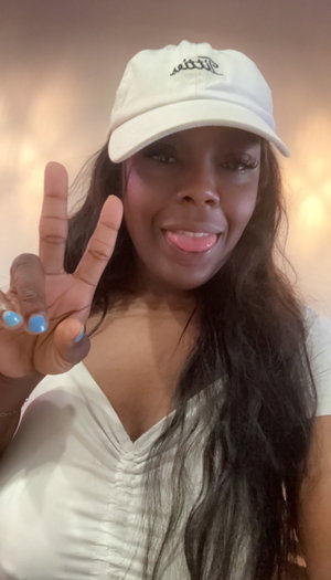 Woman with long dark hair wearing white cap and top, making peace sign indoors.