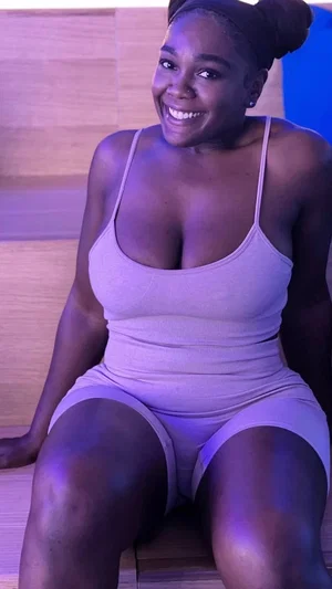 Woman in light pink bodysuit sitting indoors, smiling at camera.