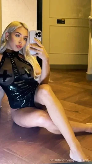Blonde woman in black latex outfit posing on wooden floor in mirror selfie.