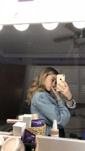 Woman with long hair in denim jacket takes mirror selfie in bedroom with makeup products visible.
