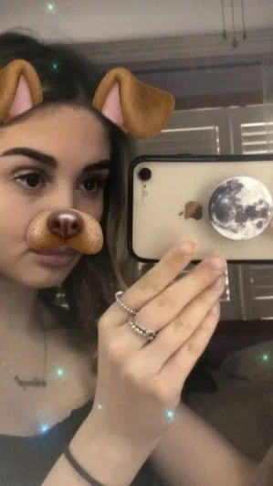 Woman with dog ears filter, holding phone, wearing dark top, indoor setting.