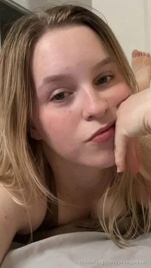 Blonde woman lying on bed, resting face on hand, wearing dark top.
