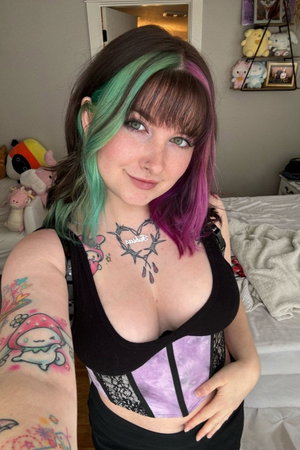 Woman with colorful hair and tattoos wearing corset in bedroom.