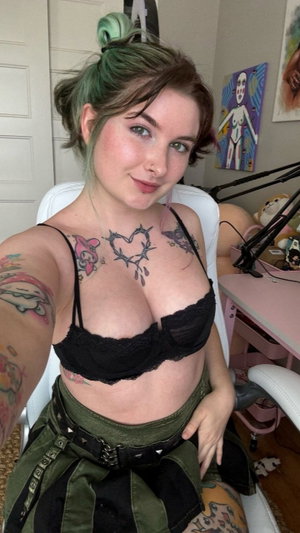 Woman with green hair and tattoos wears black lace bra and green skirt, sitting indoors.