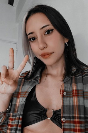 Woman with two-tone hair in open plaid shirt over black top, peace sign.