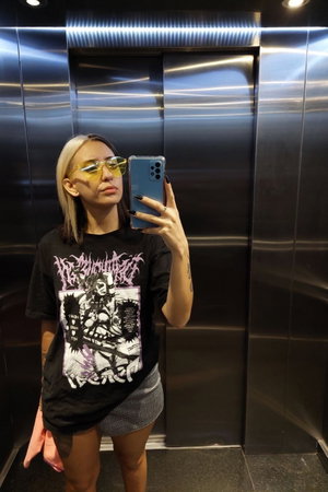 Blonde woman taking mirror selfie in elevator wearing graphic tee.