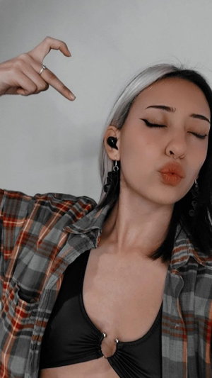 Woman with silver-tipped hair in plaid shirt over black top, posing indoors with hand gesture.