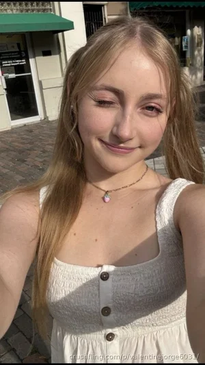 Woman with blonde hair in white sleeveless top, smiling outdoors on cobblestone street.