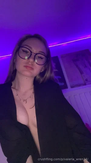 Woman with glasses and dark blazer, bare chest, in room with purple lighting.