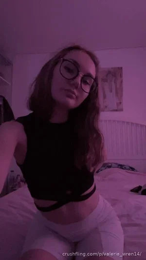Woman with glasses in black crop top and white shorts sitting on bed in dim purple light.